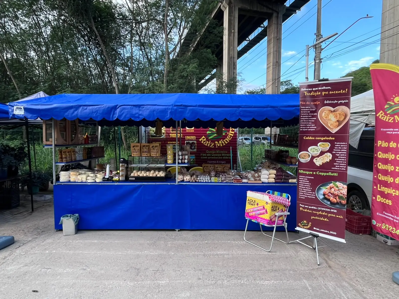 A barraca da Raiz Mineira na feira, com a tenda azul e o balcão cheio de pão de queijo, queijos e linguiças, ao lado de dois banners vermelhos. Atrás dela passam os pilares da Terceira Ponte.